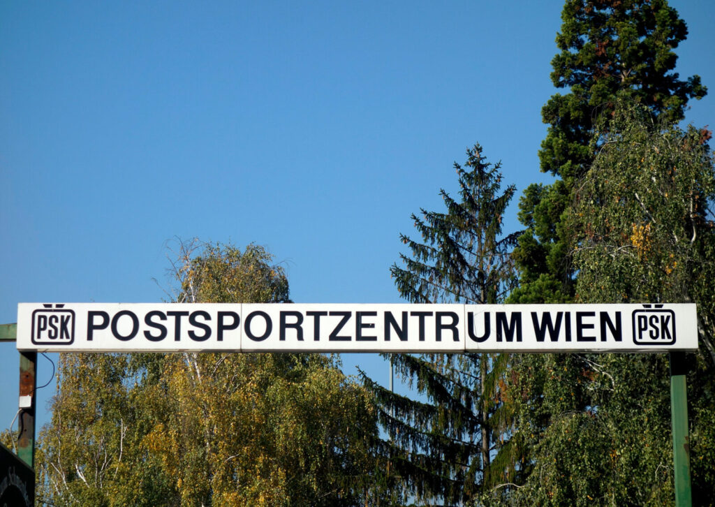 Schild mit der Aufschrift 'POSTSPORTZENTRUM WIEN' vor Bäumen und blauem Himmel