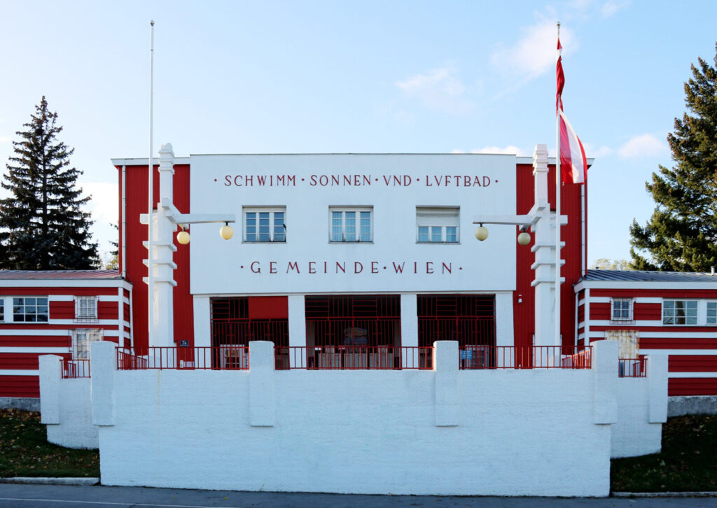 Fassade eines historischen Schwimmbads mit der Aufschrift 'SCHWIMM SONNEN UND LUFTBAD GEMEINDE WIEN' in klarer, symmetrischer Architektur und weiß-roter Farbgebung