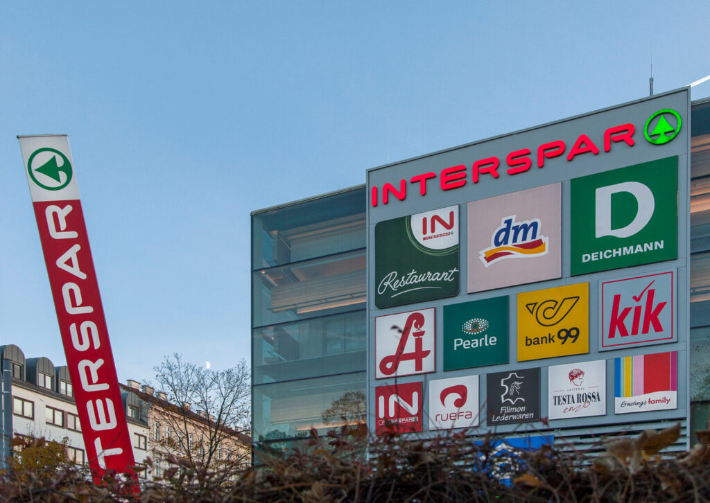 Großes Werbeschild mit Logos von INTERSPAR, dm, Deichmann, bank99, kik und weiteren Geschäften vor einem Einkaufszentrum unter blauem Himmel