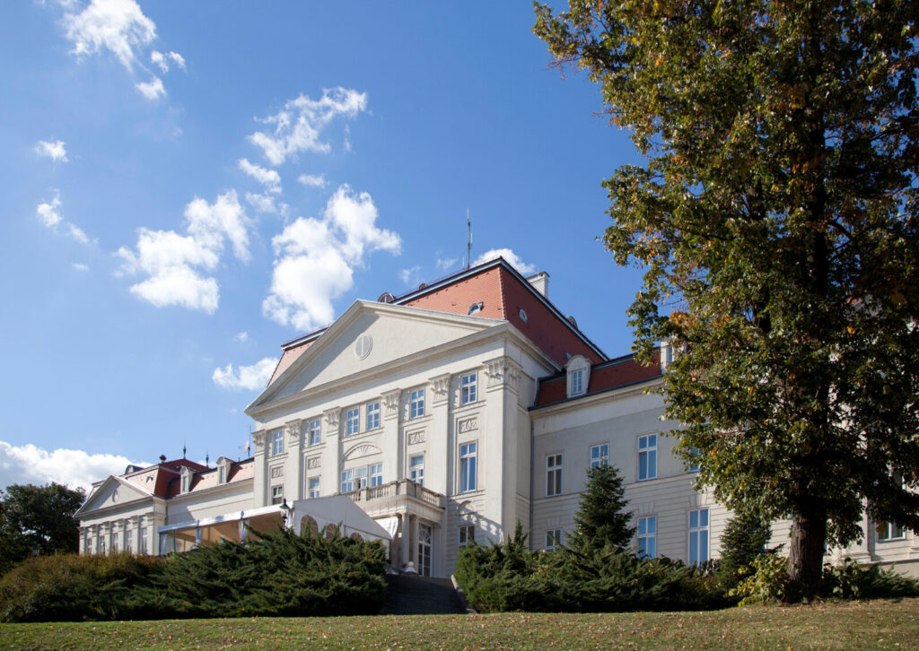 Schloss Wilhelminenberg mit klassizistischer Fassade, umgeben von Bäumen und Sträuchern unter blauem Himmel mit Wolken