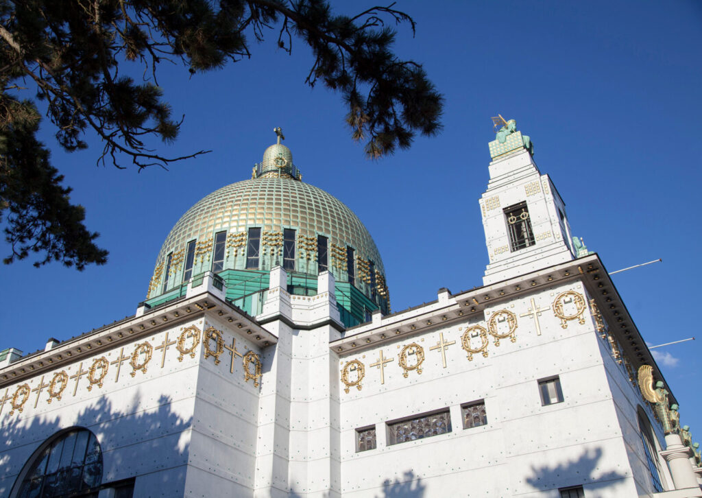 Blick auf die Kuppel und den Turm der Kirche St. Leopold mit goldenen Verzierungen vor blauem Himmel und Baumzweigen im Vordergrund